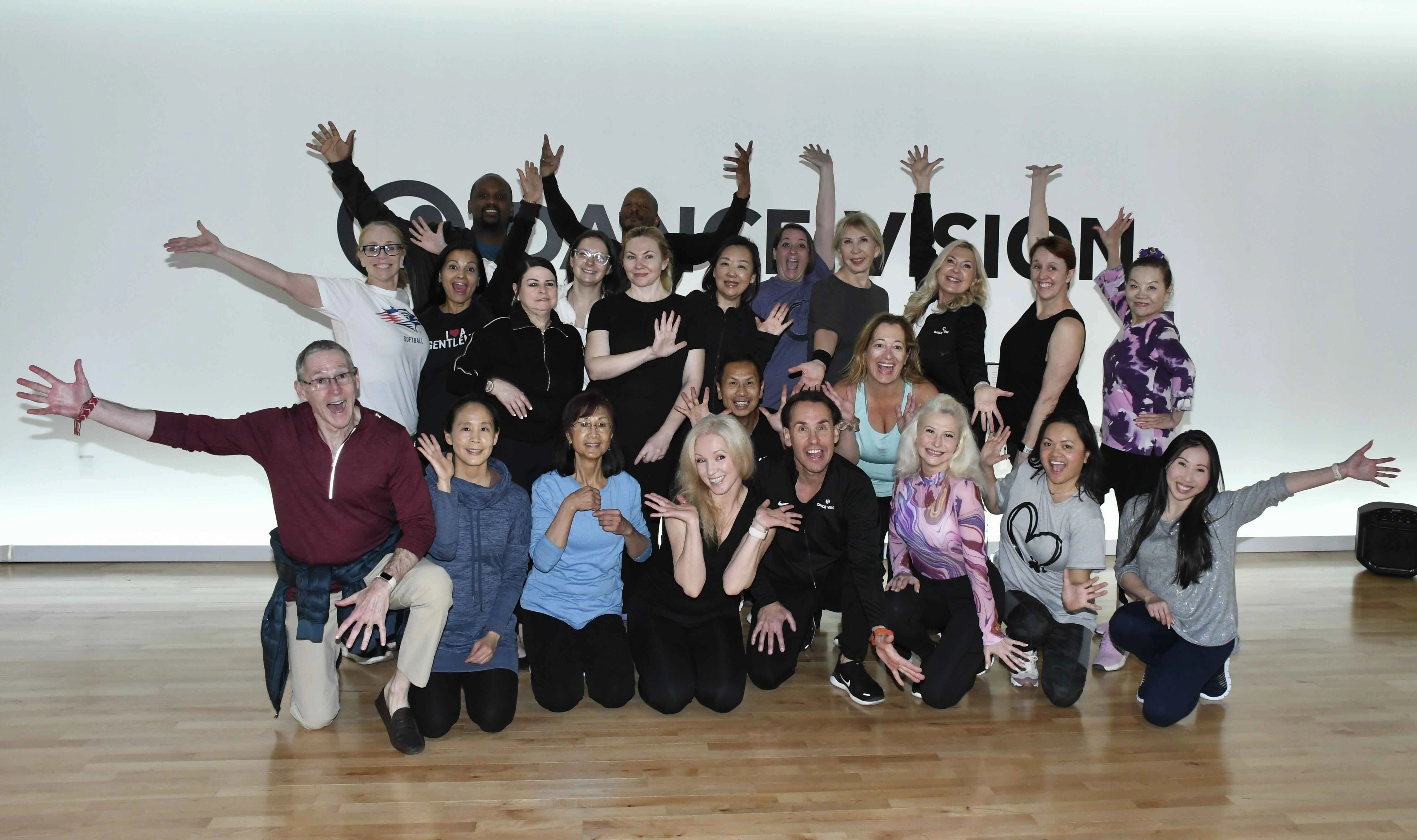 A group of people posing enthusiastically in a dance studio, smiling with arms raised.