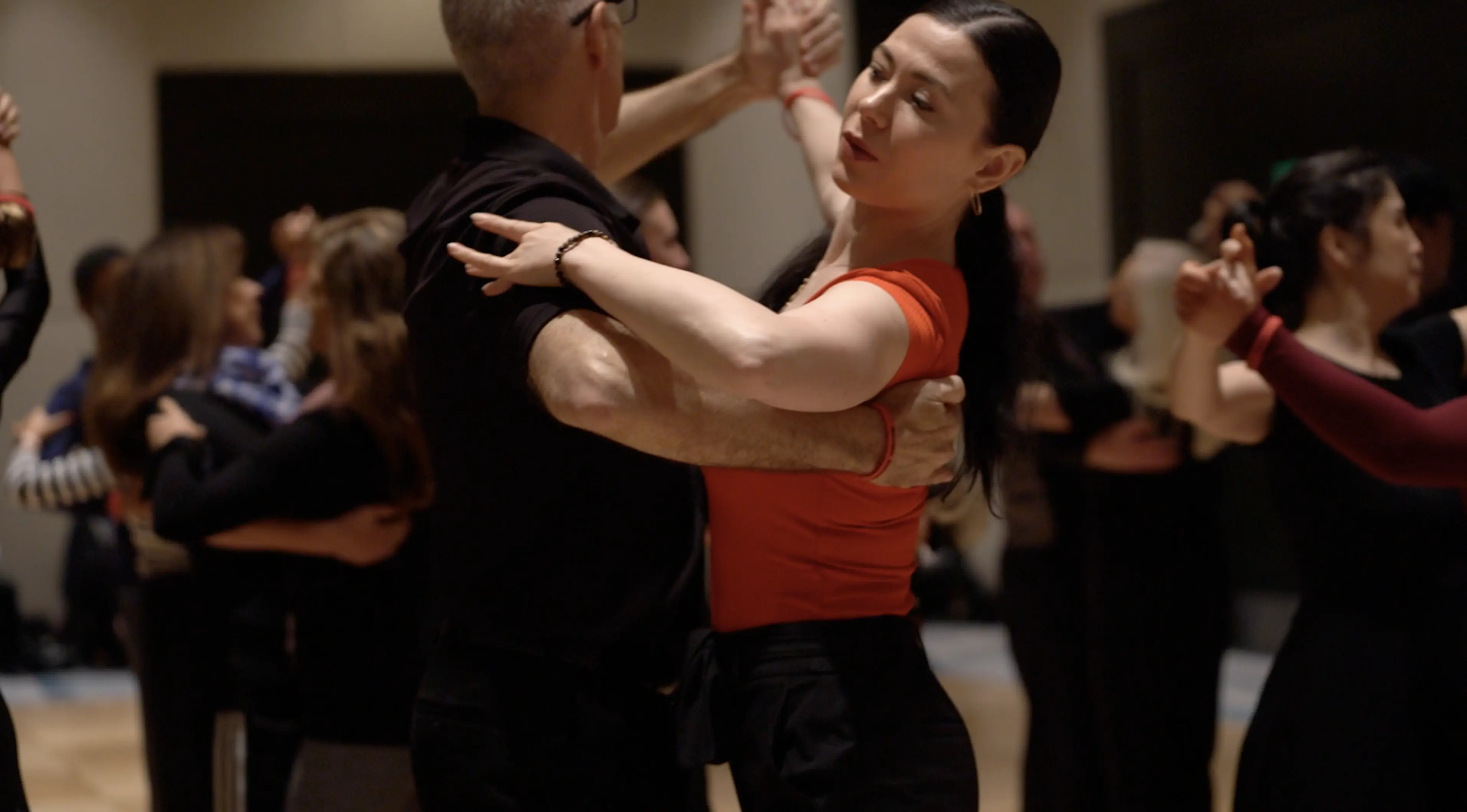 A pair dances closely in a ballroom setting, with the woman in a red top and the man in black, surrounded by other dancing pairs in the background.